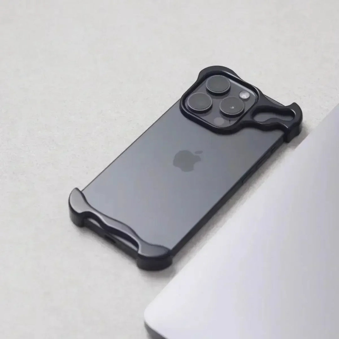 A black iPhone with a minimalist, Metal Bumper Design case is placed on a light gray surface next to the edge of a silver laptop. The case covers only the corners and camera area of the phone.
