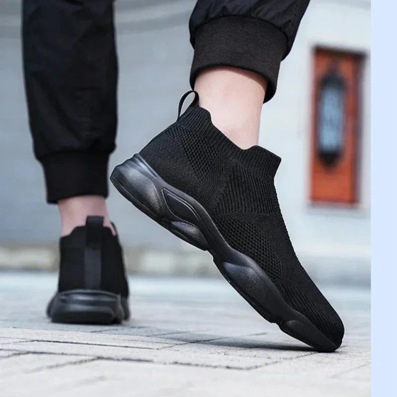 A person wearing black slip-on athletic sneakers with a non-slip sole and black pants stands on a paved surface, lifting one foot slightly off the ground. The background is blurred, keeping the focus on the shoes.