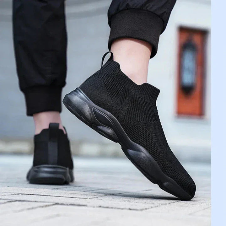 A person wearing black slip-on athletic sneakers with a non-slip sole and black pants stands on a paved surface, lifting one foot slightly off the ground. The background is blurred, keeping the focus on the shoes.