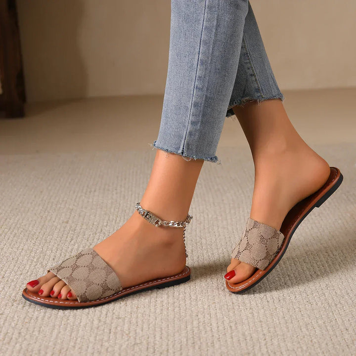 A person wearing light-wash frayed-hem jeans and beige French Sole sandals in a classic slide silhouette sits on a carpeted floor. One ankle is adorned with a silver anklet, and their toenails are painted red.