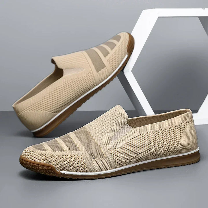 A pair of beige men's casual loafers with brown rubber soles, white accents, and canvas lining are displayed against a gray background, with one breathable shoe propped upright on a geometric white frame.