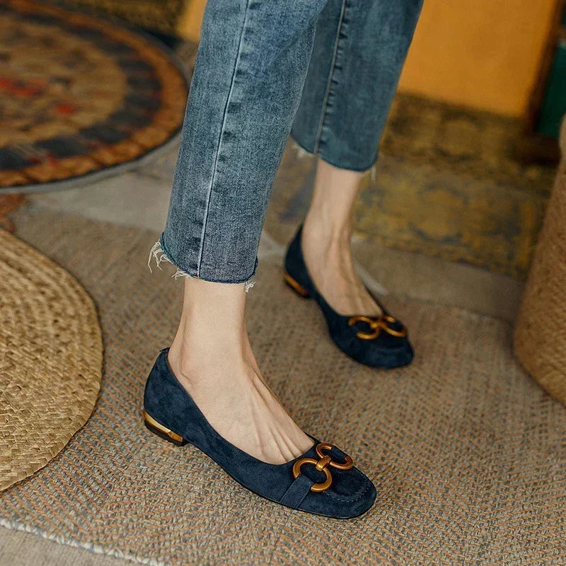 A person wearing frayed-hem skinny jeans and navy blue suede flats with cushioned insoles and gold decorative buckles stands on a woven rug in a cozy room with patterned rugs and warm-toned decor.