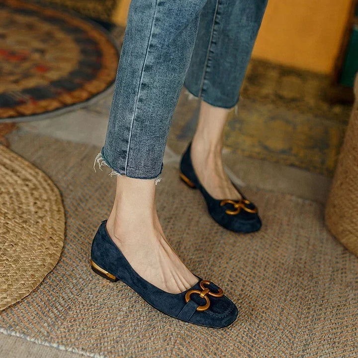 A person wearing frayed-hem skinny jeans and navy blue suede flats with cushioned insoles and gold decorative buckles stands on a woven rug in a cozy room with patterned rugs and warm-toned decor.