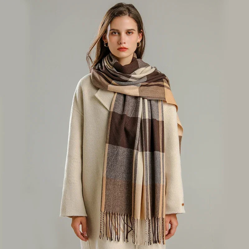 A woman with long brown hair wears a cream-colored coat and a large, lightweight plaid scarf in shades of brown, beige, and gray. She stands against a plain, light gray background, looking at the camera.