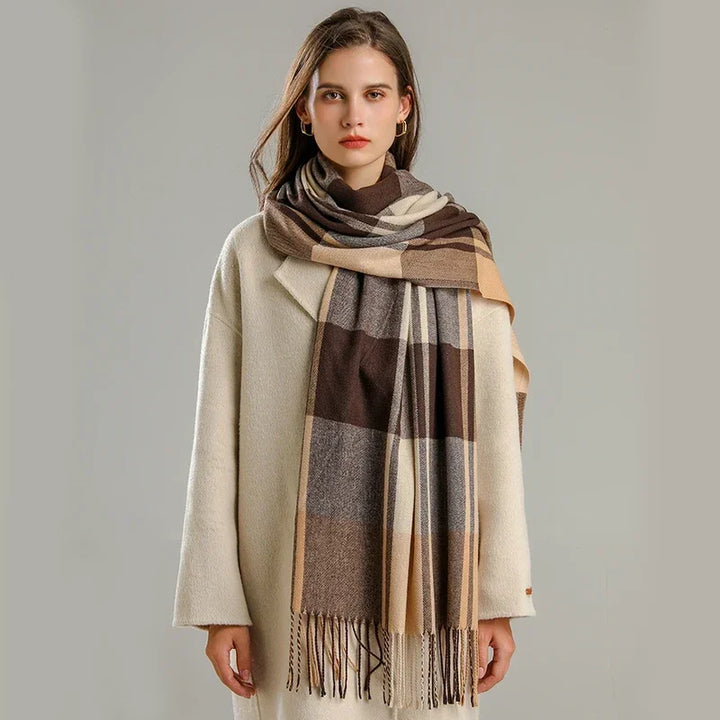 A woman with long brown hair wears a cream-colored coat and a large, lightweight plaid scarf in shades of brown, beige, and gray. She stands against a plain, light gray background, looking at the camera.