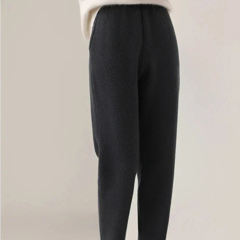 A person wears high-waisted, loose-fitting black pants with a slim tailored leg and ultra soft insulation, paired with a white, long-sleeve top. The background is plain and neutral. The upper body and face are not visible.