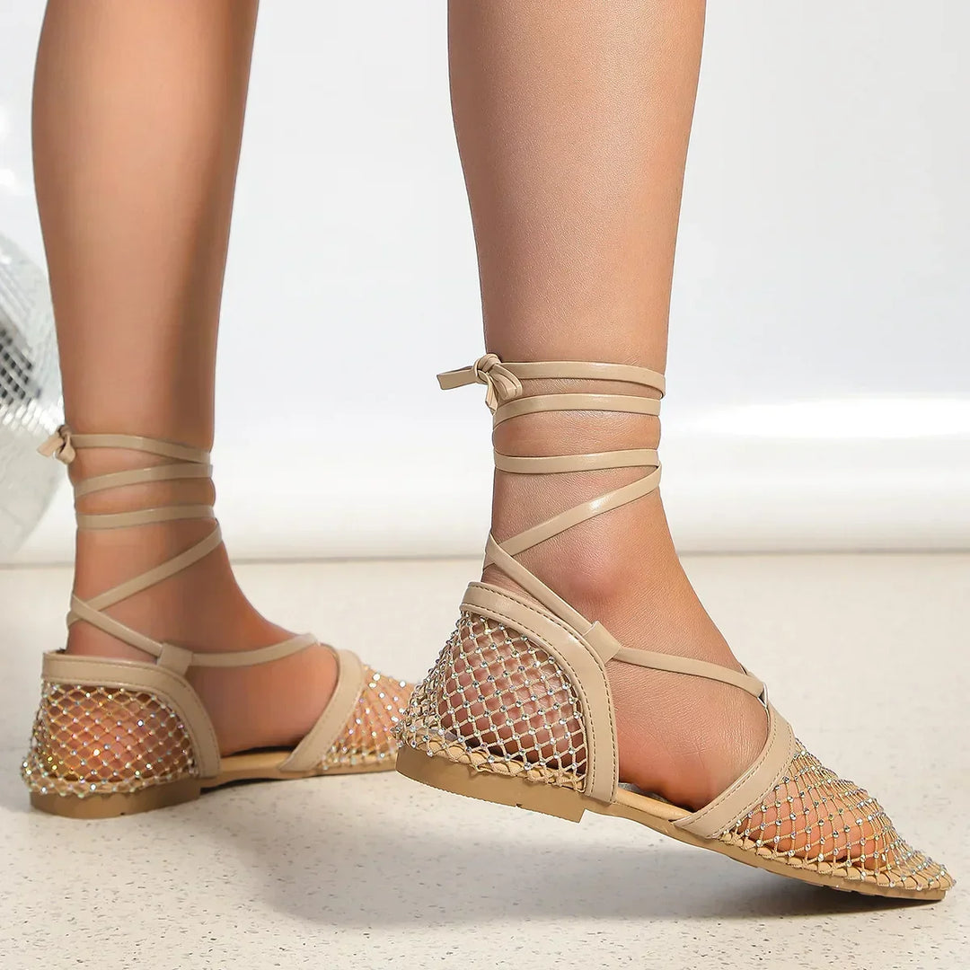 A person wearing beige flat sandals with a mesh upper and ankle wrap straps, featuring slip-resistant soles, stands on a light-colored floor.