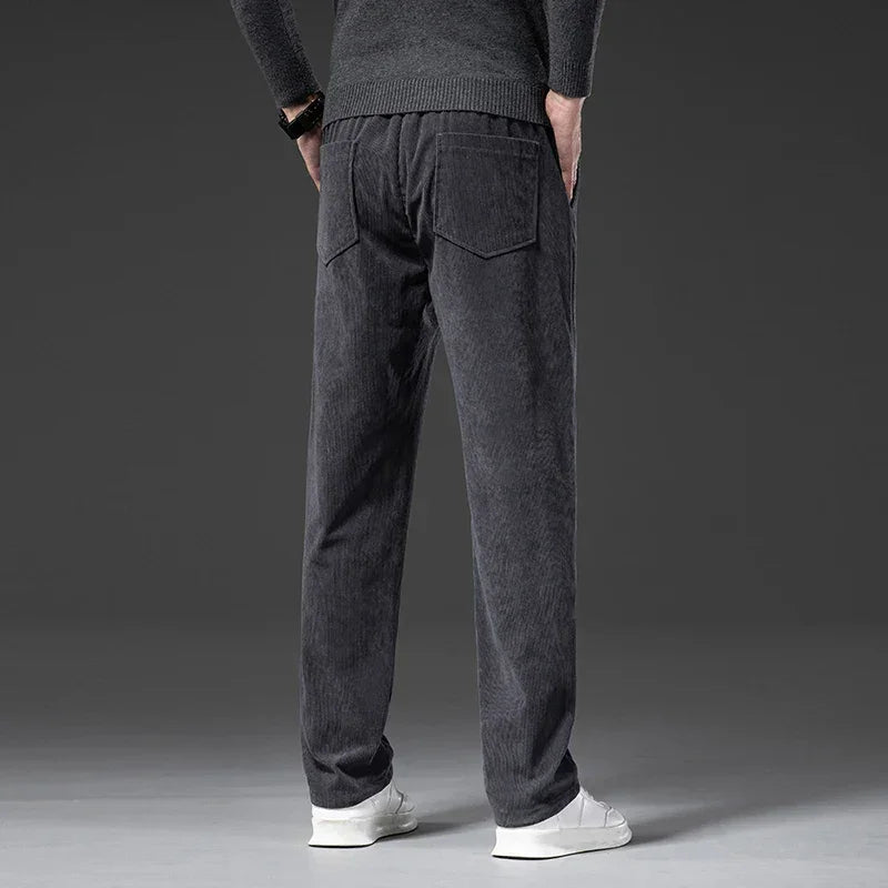 A person wearing a dark sweater, men's smart casual pants in dark gray corduroy with a zipper fly, and white sneakers stands facing away, hands behind their back, against a plain gray background.