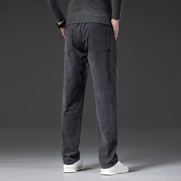 A person wearing a dark sweater, men's smart casual pants in dark gray corduroy with a zipper fly, and white sneakers stands facing away, hands behind their back, against a plain gray background.