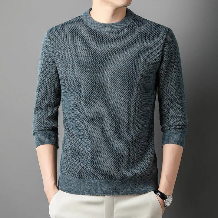 A person wears a relaxed fit sweater in textured teal with long sleeves and light-colored pants, standing against a gray background with hands in pockets. Their face is partially out of frame.