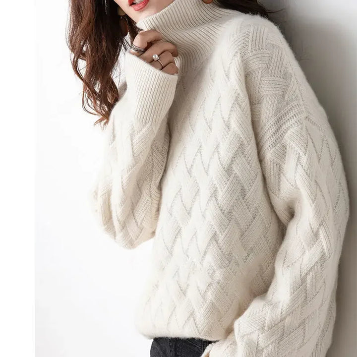 A person wearing a cozy, ivory-colored, ultra soft cable knit turtleneck sweater with long sleeves and textured patterns, paired with dark pants. The persons face is partially obscured by their hand.