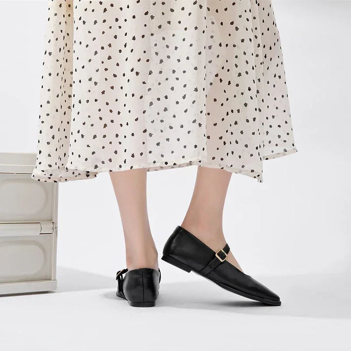 A person wearing a white, polka-dotted skirt and black Mary Jane shoes stands next to a light-colored box, with only their legs and feet visible on a white background.