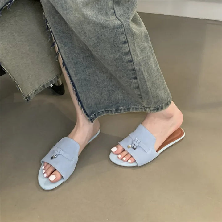 A person wearing frayed-hem gray jeans and light blue open-toe slide sandals with a suede finish and silver buckle details, showing white-painted toenails, stands on a smooth beige floor.
