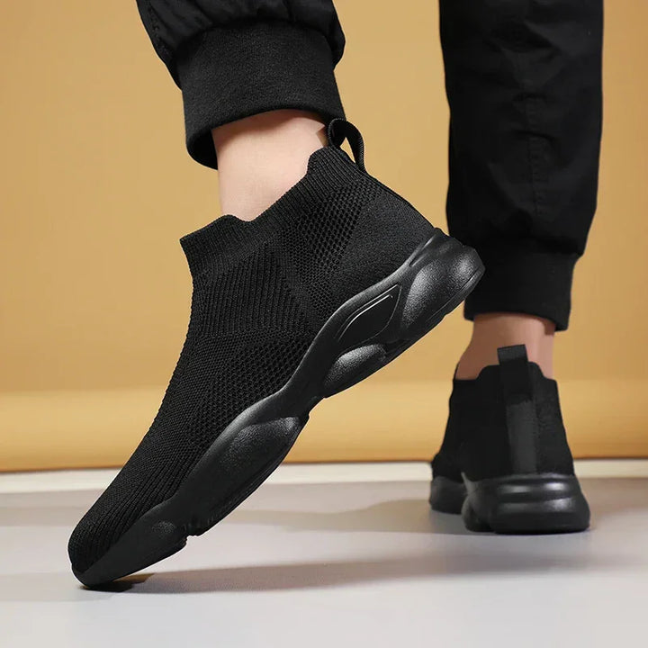 A person wearing black slip-on athletic sneakers with breathable knit fabric and thick, non-slip soles stands with one foot slightly raised against a neutral beige background.