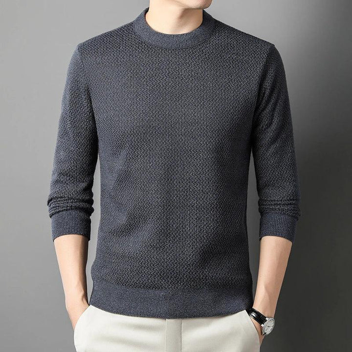 A person wearing a high-quality cotton sweater in dark gray with long sleeves and light-colored pants stands against a plain gray background, hands in pockets. The persons head is not fully visible.