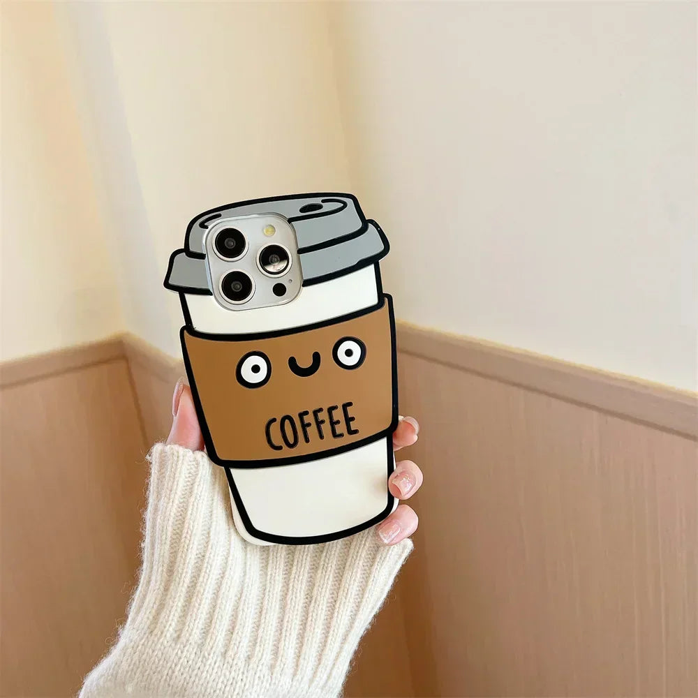 A hand in a white sweater holds a phone with a 3D coffee cup case, made of soft silicone for shockproof protection. The case features a smiling cartoon cup with a brown sleeve labeled COFFEE against a light wall and wood paneling.