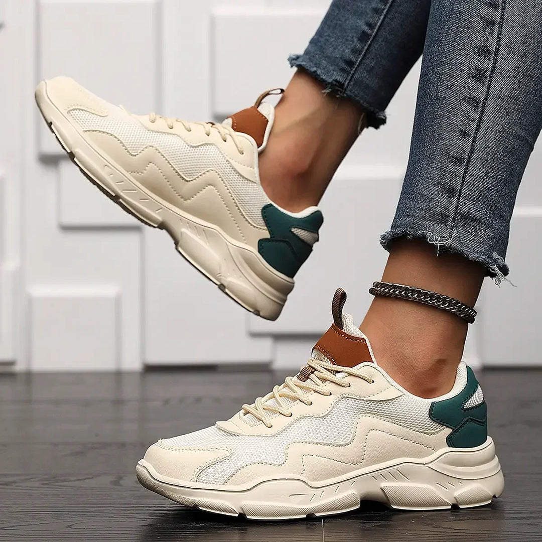 A person wearing cream-colored slip-on street shoes with green and brown accents, paired with frayed-hem skinny jeans and a silver anklet, stands on a dark wooden floor with a white textured wall in the background.