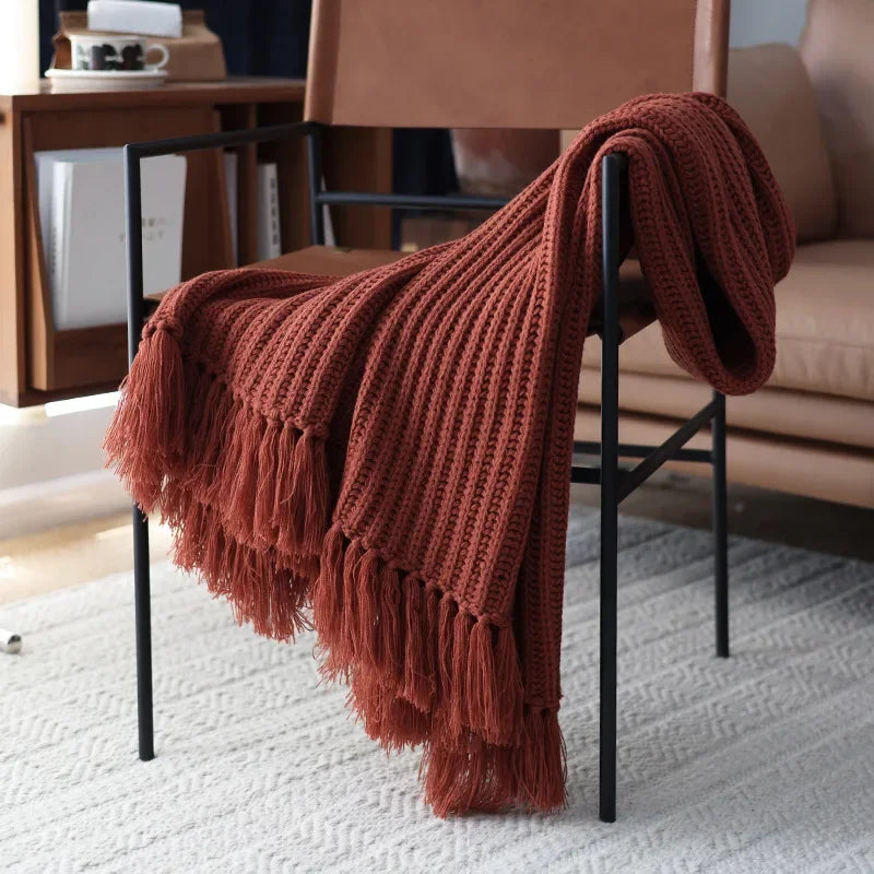 A rust-colored chunky weave throw with thick tassels drapes over a modern black metal chair, placed on a light textured rug near a tan leather sofa in a cozy living room setting.