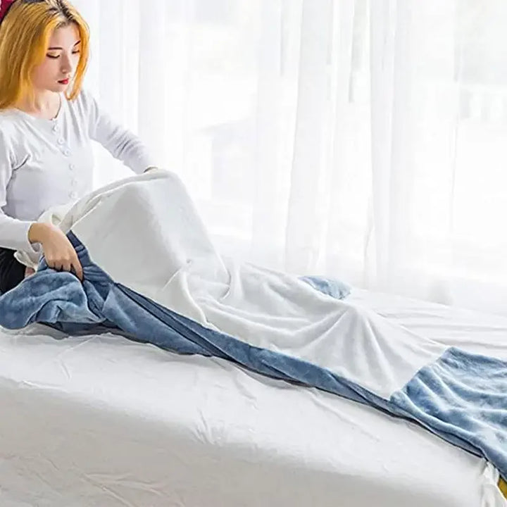 A person with blonde hair, wearing a white long-sleeve top and cozy pajamas, sits on a bed and covers their legs with a blue and white mermaid tail blanket. Light streams in through sheer curtains behind them.