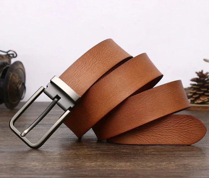A brown cowhide belt with a silver buckle is coiled on a wooden surface. A pine cone and a vintage camera are blurred in the background.
