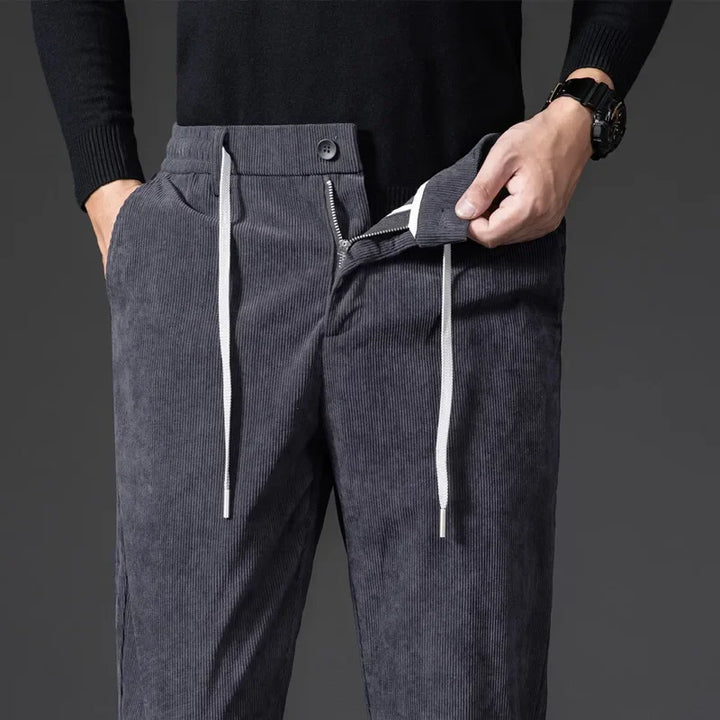 A person wearing a black sweater and men's smart casual corduroy pants with white drawstrings and a zipper fly stands with one hand in their pocket and the other adjusting the waistband.