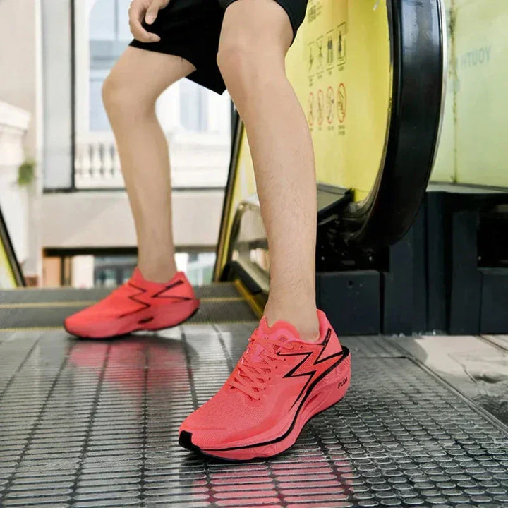 A person wearing bright pink running shoes with a non-slip outsole stands on an escalator, shown from the knees down. The individual is dressed in black shorts.
