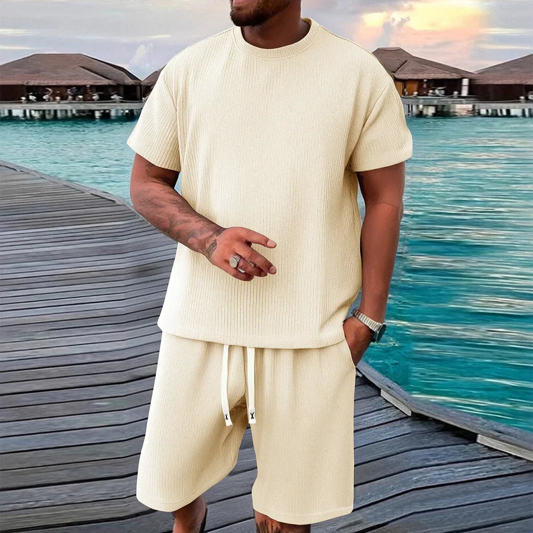 Man wearing beige short sleeve knit set on boardwalk near water villas at sunset, Rovelli fashion.