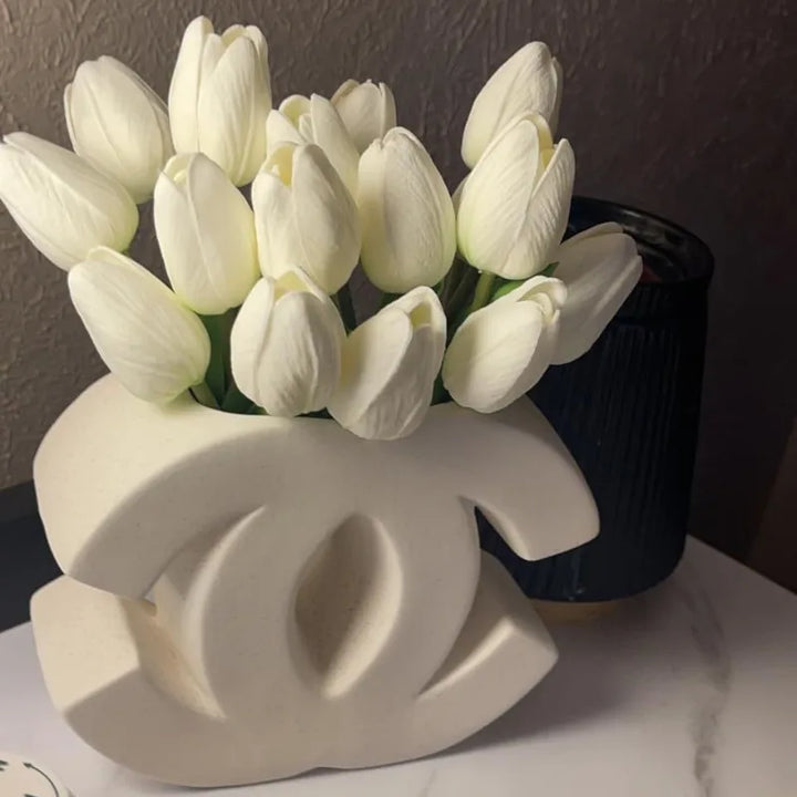 A Ceramic Vase shaped like the Chanel logo holds white tulips, placed on a white surface next to a dark, ribbed container against a textured wall—perfect for Nordic Home Decoration or Irregular INS Style interiors.