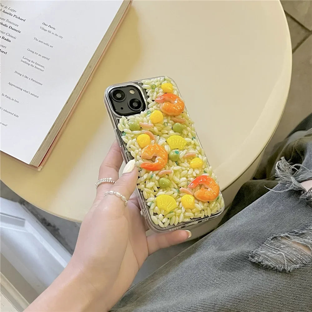 A hand holds a phone with a case designed to look like fried rice, featuring realistic 3D detailing—shrimp, corn, green peas, and egg. The person sits at a round table with an open book and wears ripped jeans.