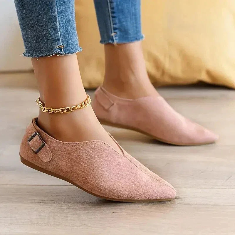 A person wearing light pink pointed toe shoes with a side buckle and gold chain anklet, featuring a cushioned insole, paired with frayed hem blue skinny jeans, standing on a wooden floor.