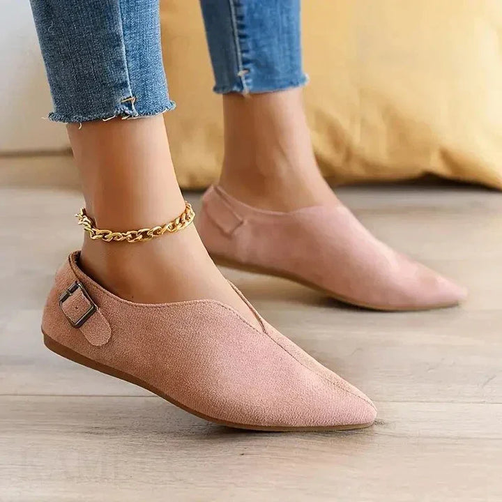 A person wearing light pink pointed toe shoes with a side buckle and gold chain anklet, featuring a cushioned insole, paired with frayed hem blue skinny jeans, standing on a wooden floor.