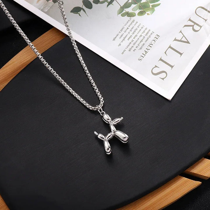 A playful balloon dog necklace pendant hangs from an adjustable chain necklace, displayed on a black surface with a wooden accent and part of an open magazine in the background.
