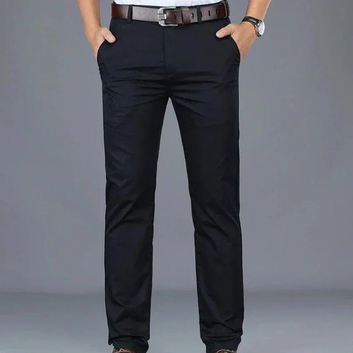 A person wearing dark blue dress pants featuring a tailored fit and hands in pockets, a brown belt, a gray shirt tucked in, and a wristwatch, standing against a plain gray background.