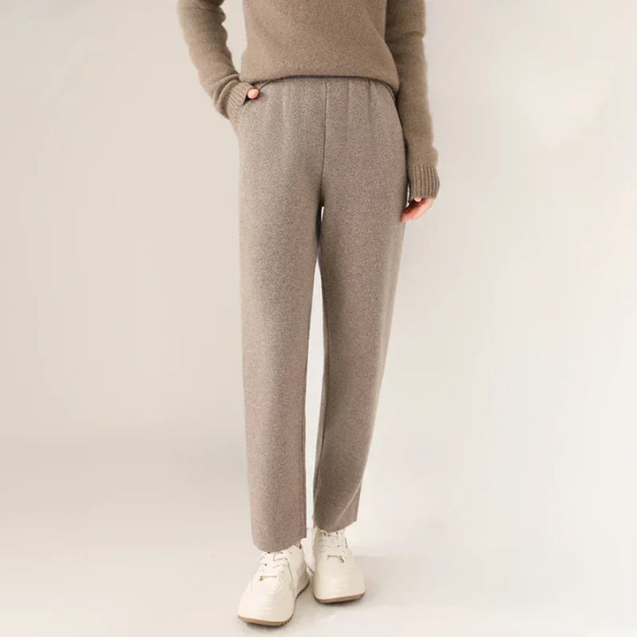 A person wears a taupe sweater and matching slim tailored leg trousers with white sneakers, standing against a plain light background with hands in pockets.