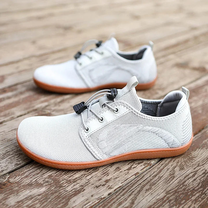 A pair of light gray casual sneakers with breathable mesh uppers, a wide toe box, gray laces, and brown cushioned soles are placed on a wooden surface. One shoe is in the foreground, the other slightly out of focus in the background.