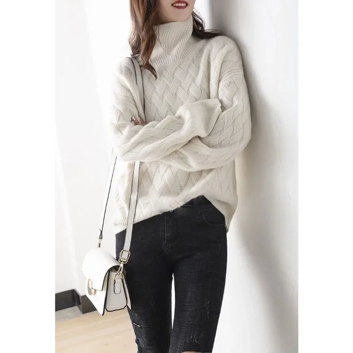 A woman leans against a white wall, wearing an ultra soft, cream-colored turtleneck sweater with a textured knit pattern, black ripped jeans, and carrying a small white shoulder bag. Her face is partially turned away.