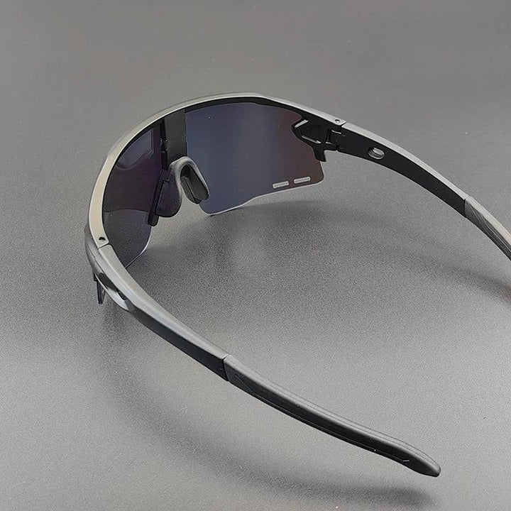 A pair of black sports sunglasses with dark, semi-rimless polarized lenses is placed on a smooth, gray surface. The arms and nose pads are visible, and the frame has a modern, angular design.