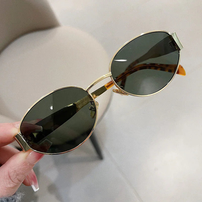A hand with long, manicured nails holds vintage-inspired sunglasses with gold frames and dark lenses over a light-colored floor and a beige chair in the background.