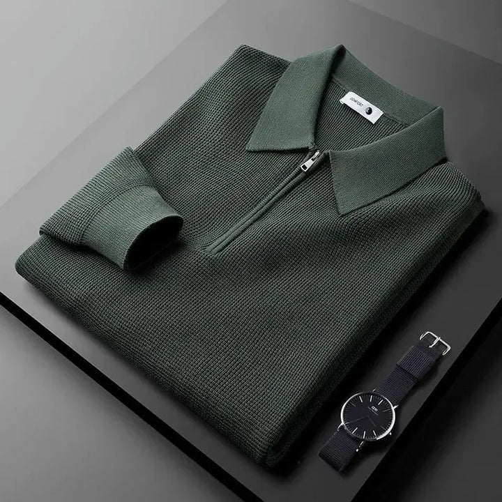 A folded dark green zip-collar long sleeve polo in textured, stretchy fabric lies on a dark surface next to a minimalist black wristwatch with a fabric strap.