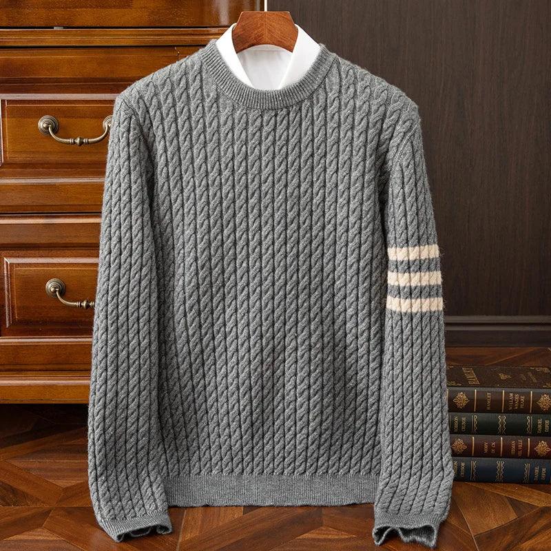 A gray cable-knit cashmere sweater with three white stripes on one sleeve is displayed on a wooden hanger in front of a brown wooden dresser and a stack of books. A white shirt is layered underneath the sweater.