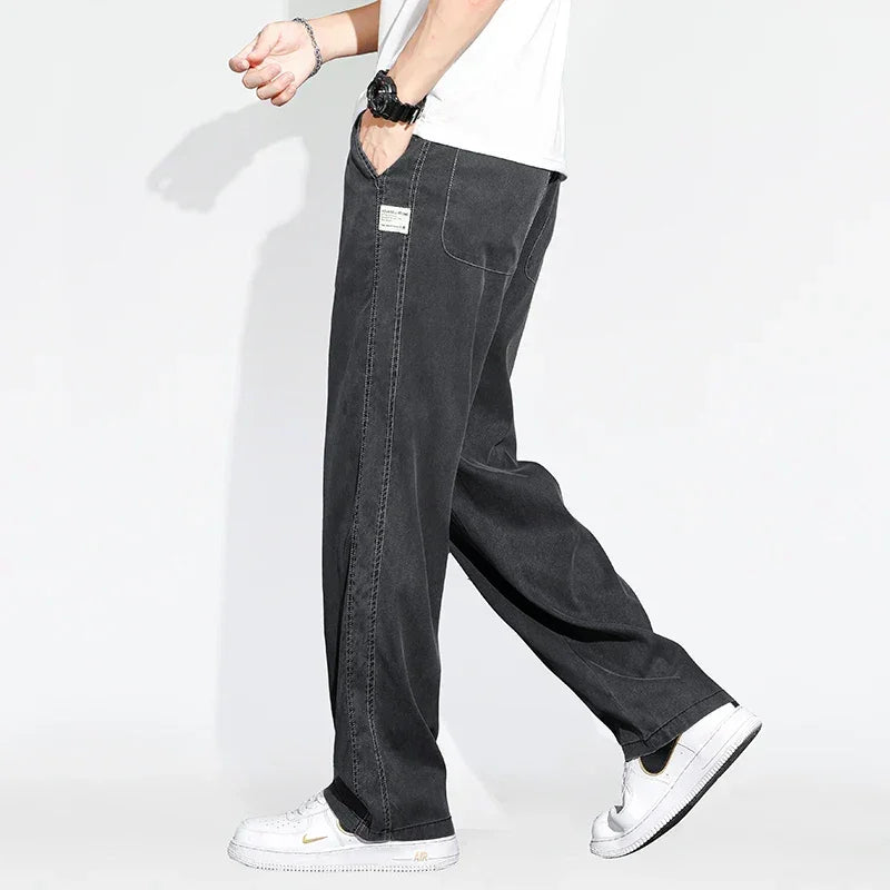 A person wears Summer Loose Men Jeans—wide-leg denim trousers with visible stitching—paired with a white T-shirt, black wristwatch, and white sneakers against a plain white background. Only the lower body is shown.