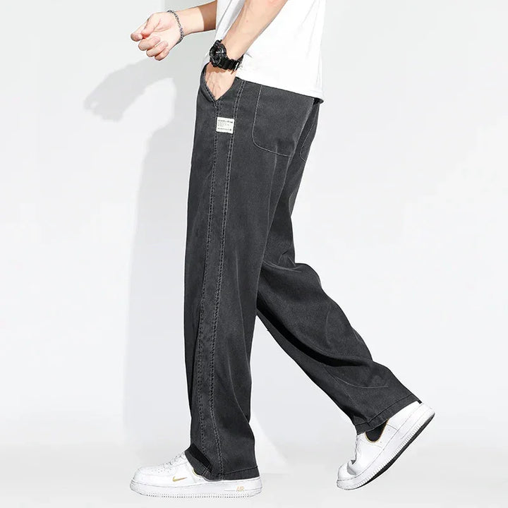 A person wears Summer Loose Men Jeans—wide-leg denim trousers with visible stitching—paired with a white T-shirt, black wristwatch, and white sneakers against a plain white background. Only the lower body is shown.