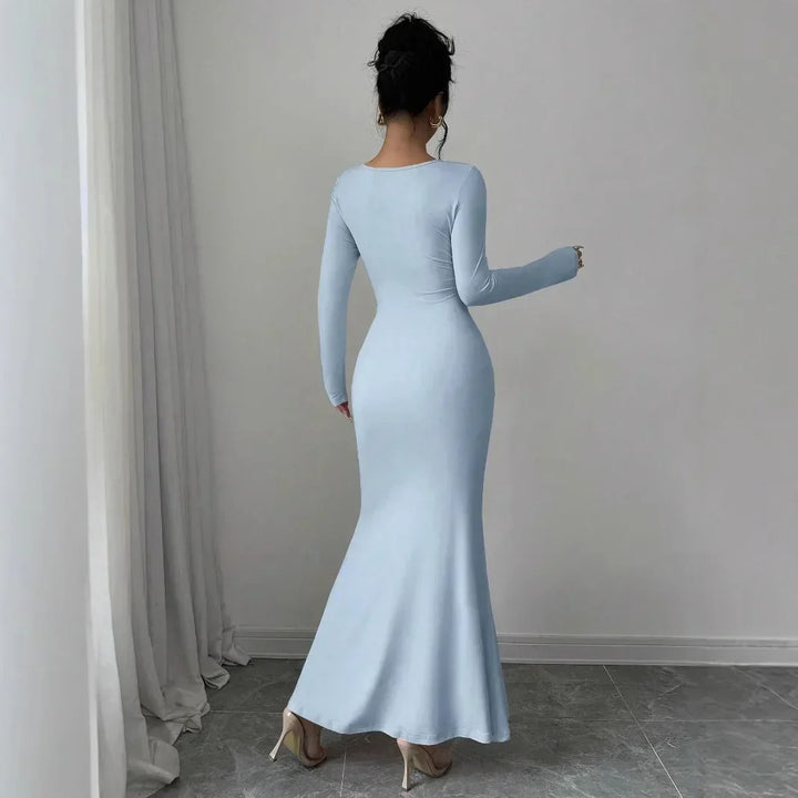 Woman in long-sleeve light blue bodycon maxi dress, standing in modern minimalist room, Rovelli fashion