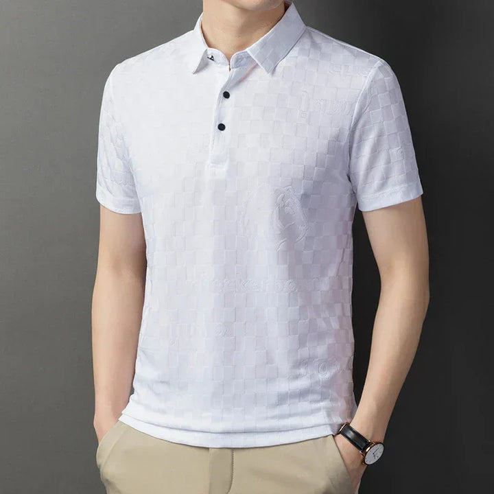 A man wears a tailored fit, short-sleeve white polo shirt with a subtle checkerboard pattern, three black buttons, and beige pants. His hands are in his pockets, and he sports a black watch against a gray background.