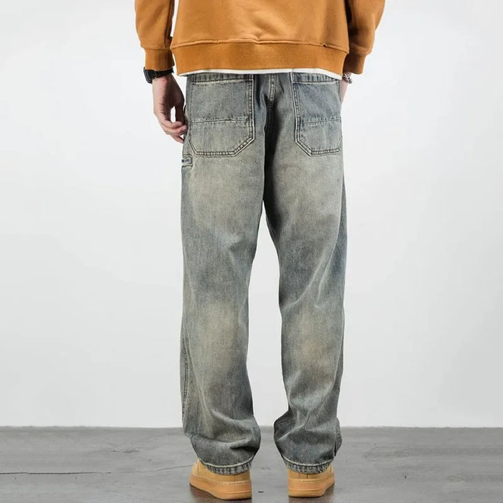 A person stands facing away, wearing a brown sweatshirt, loose-fitting stone washed jeans, and tan shoes, against a plain light background.