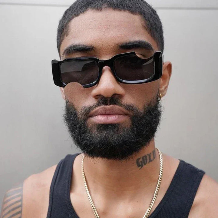 A man with a short beard and mustache wears avant-garde sunglasses with UV400 protection, a black tank top, a gold chain, and has a tattoo reading GOD on his neck. He stands in front of a plain, light-colored background.