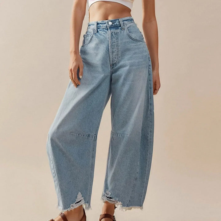 A person wearing high-waisted, relaxed fit light blue jeans with frayed hems and a white crop top, standing against a plain beige background.