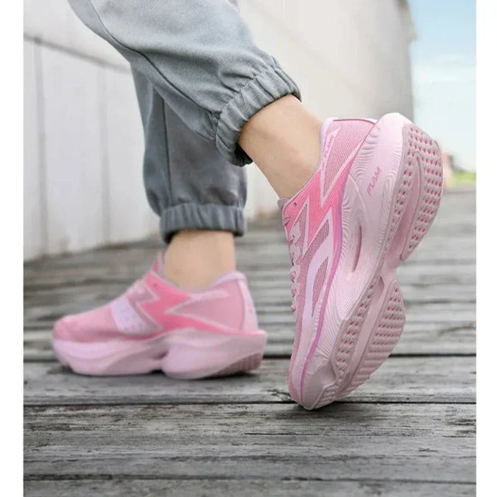 Person wearing ultra-lightweight light pink sneakers with thick, shock-absorbing soles and light gray jogger pants walks on a wooden pathway beside a white wall. The photo focuses on the shoes and lower legs.