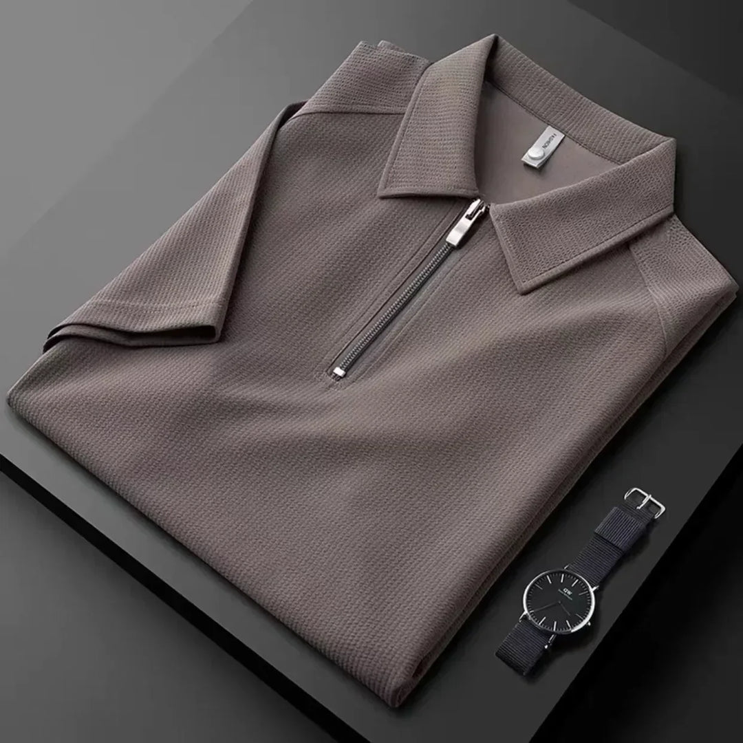 A neatly folded taupe mens silk polo shirt with a zip-up collar is displayed on a dark surface next to a minimalist black wristwatch with a silver case and black strap.