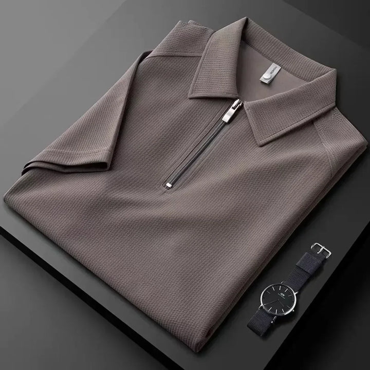 A neatly folded taupe mens silk polo shirt with a zip-up collar is displayed on a dark surface next to a minimalist black wristwatch with a silver case and black strap.
